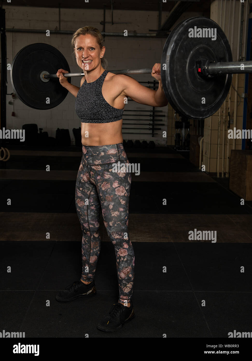 A beautiful middle aged blonde woman with Sixpack is doing back squats with the barbell. Functional fitness and weight lifting workout in a gym. Stock Photo