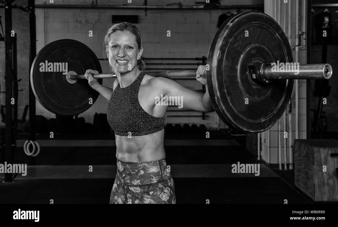 A beautiful middle aged blonde woman with Sixpack is doing back squats with the barbell. Functional fitness and weight lifting workout in a gym. Stock Photo