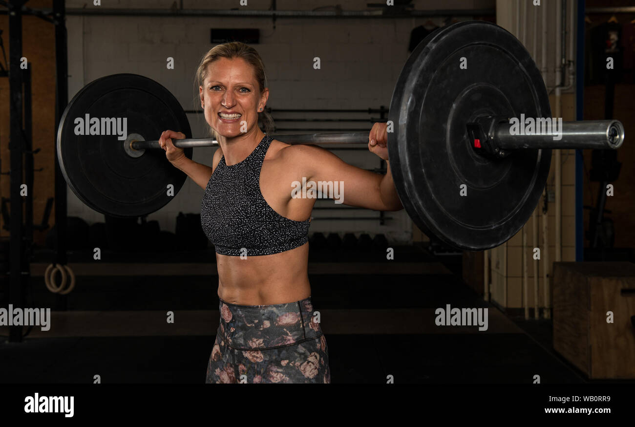 A beautiful middle aged blonde woman with strong abs is doing back squats with the barbell. Functional fitness and weight lifting workout in a gym. Stock Photo