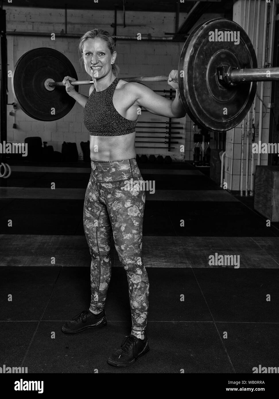 A beautiful middle aged blonde woman with strong abs is doing a clean with the barbell. Functional fitness and weight lifting workout in a gym. Stock Photo