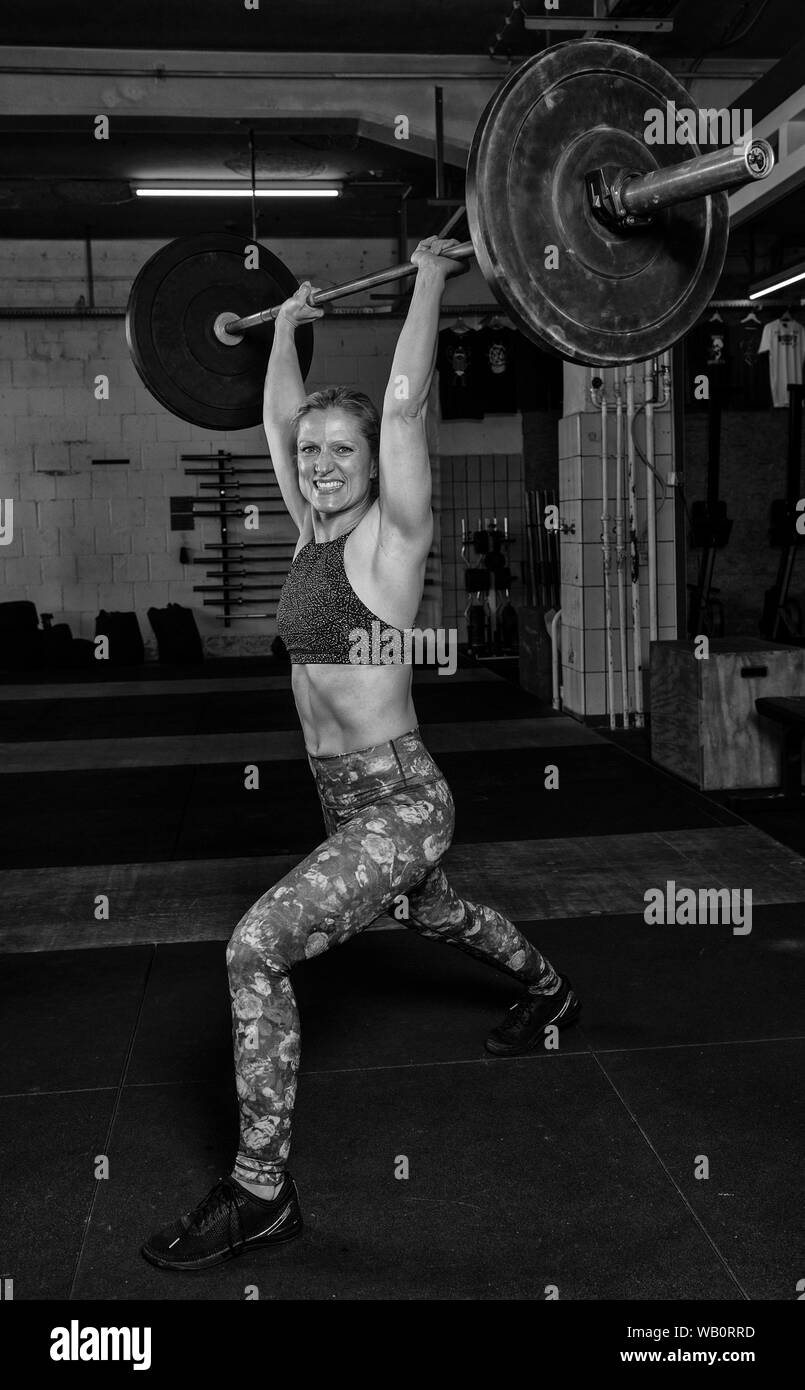 An attractive older blonde woman with Sixpack is doing a split jerk with the barbell. Functional fitness and weight lifting workout in a gym. Stock Photo
