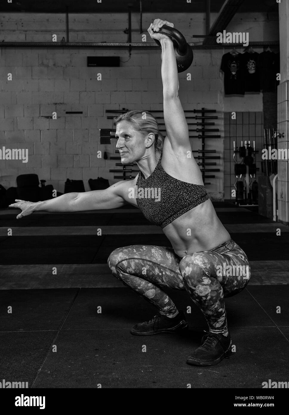 An atttractive middle aged  woman with Sixpack is doing overhead squats with a kettlebell. Functional fitness and weight lifting workout in a gym. Stock Photo