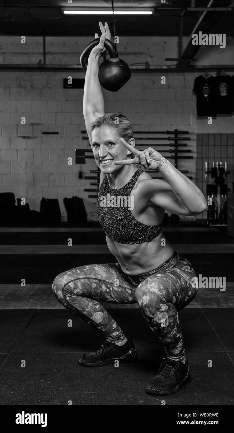 An handsome middle aged strong woman with Sixpack is doing overhead squats with a kettlebell. Functional fitness and weight lifting workout in a gym. Stock Photo