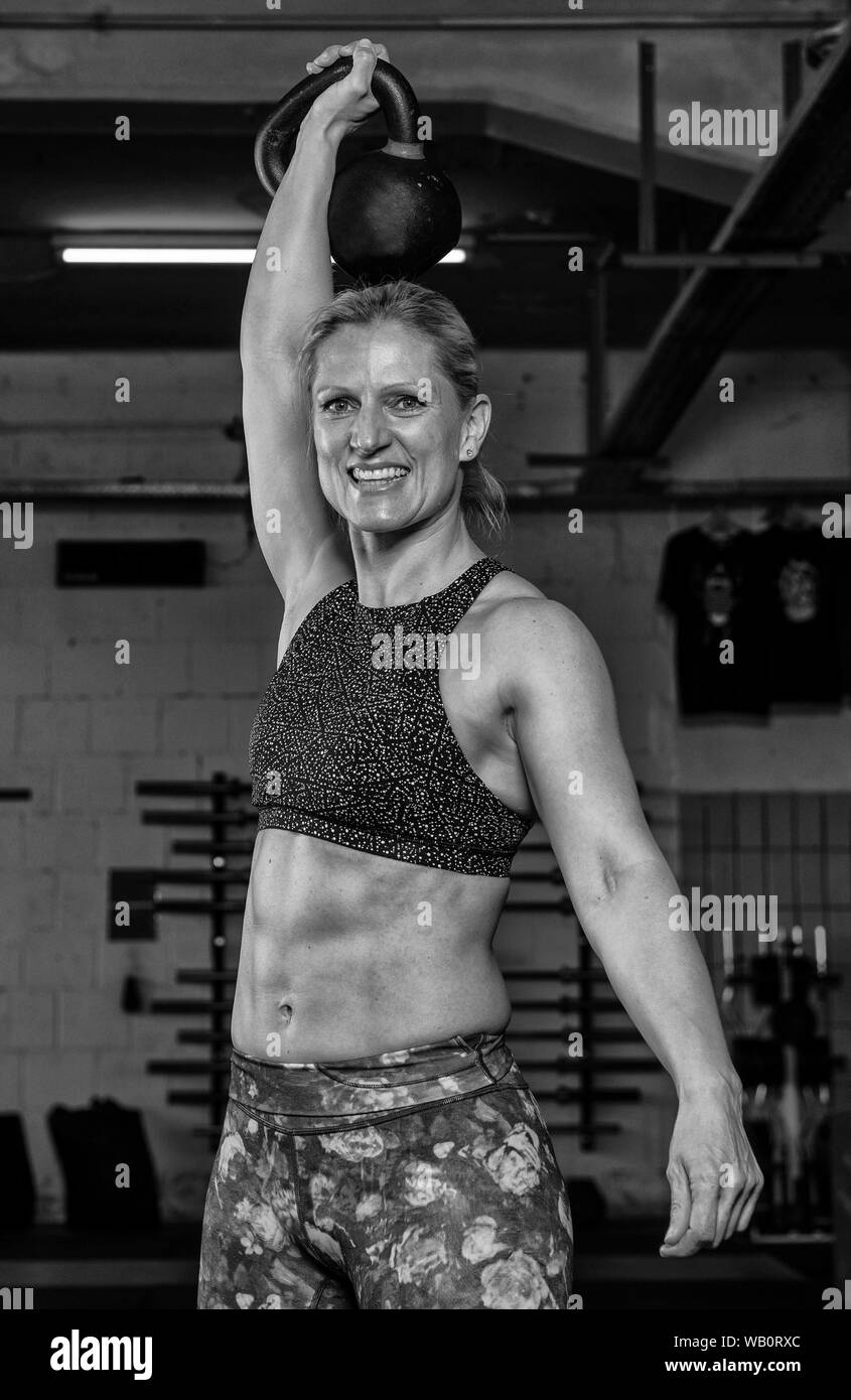 An attractive older strong woman with Sixpack is doing overhead squats with a kettlebell. Functional fitness and weight lifting workout in a gym. Stock Photo