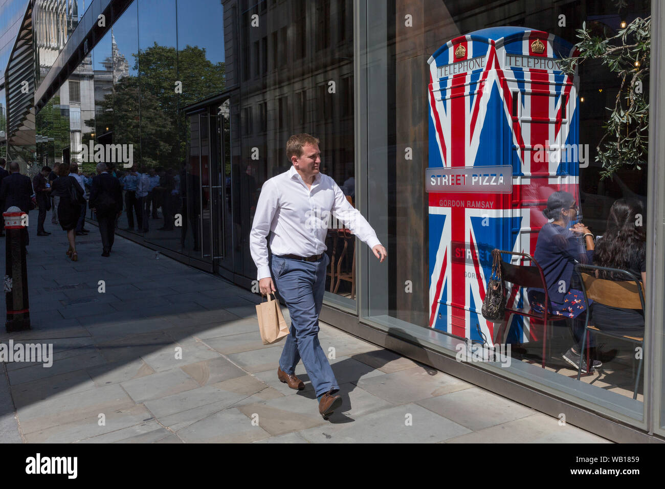 Lunchtime City workers walk past the Union Jack-themes telephone kiosk in celebrity chef Gordon Ramsey's restaurant Bread Street Kitchen on (the former Roman thoroughfare) Watling Street, in the City of London, the capital's financial district (aka the Square Mile), on 22nd August 2019, in London, England. Stock Photo