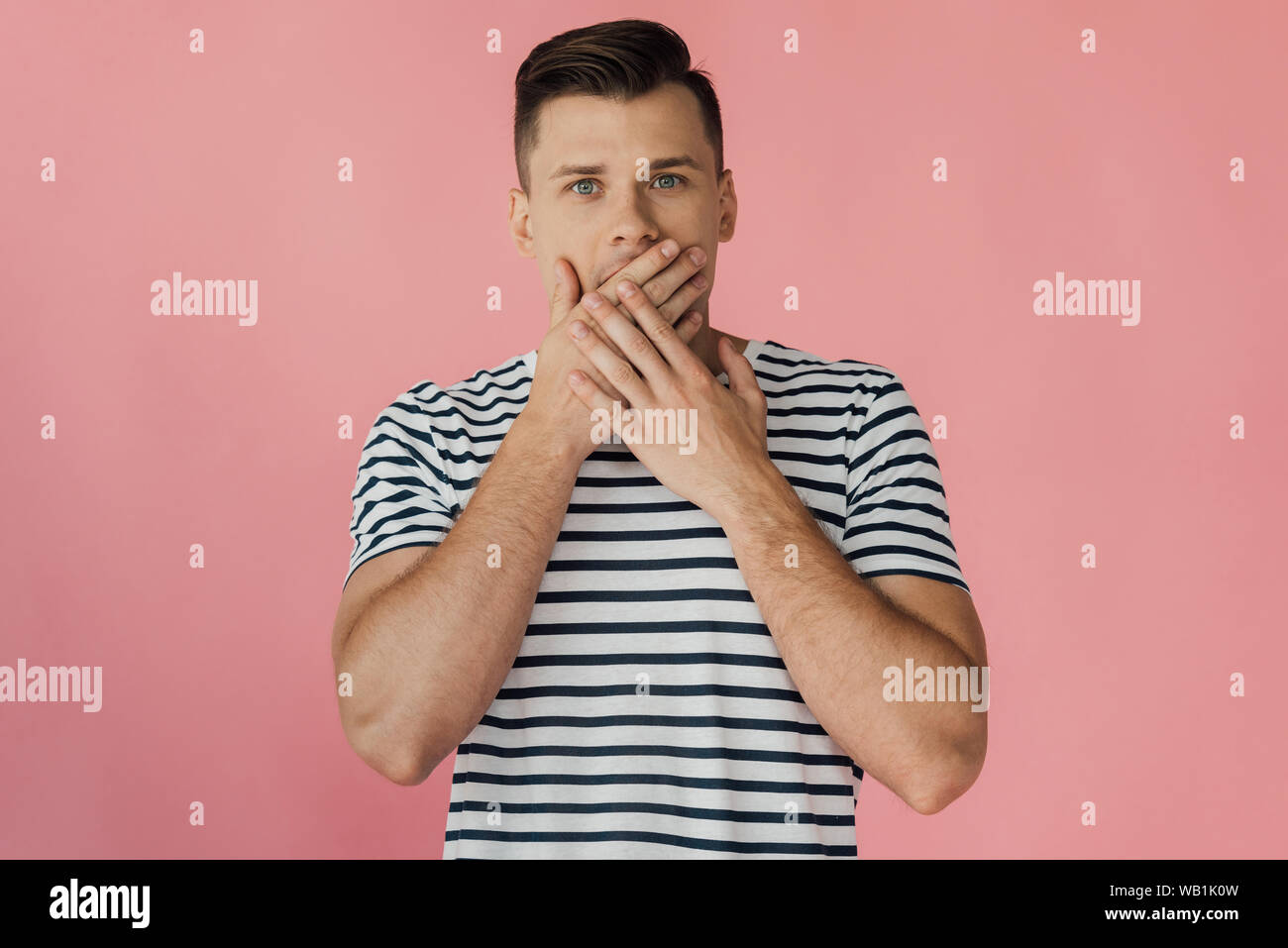 front view of shocked young man in striped t-shirt covering mouth with hands isolated on pink Stock Photo