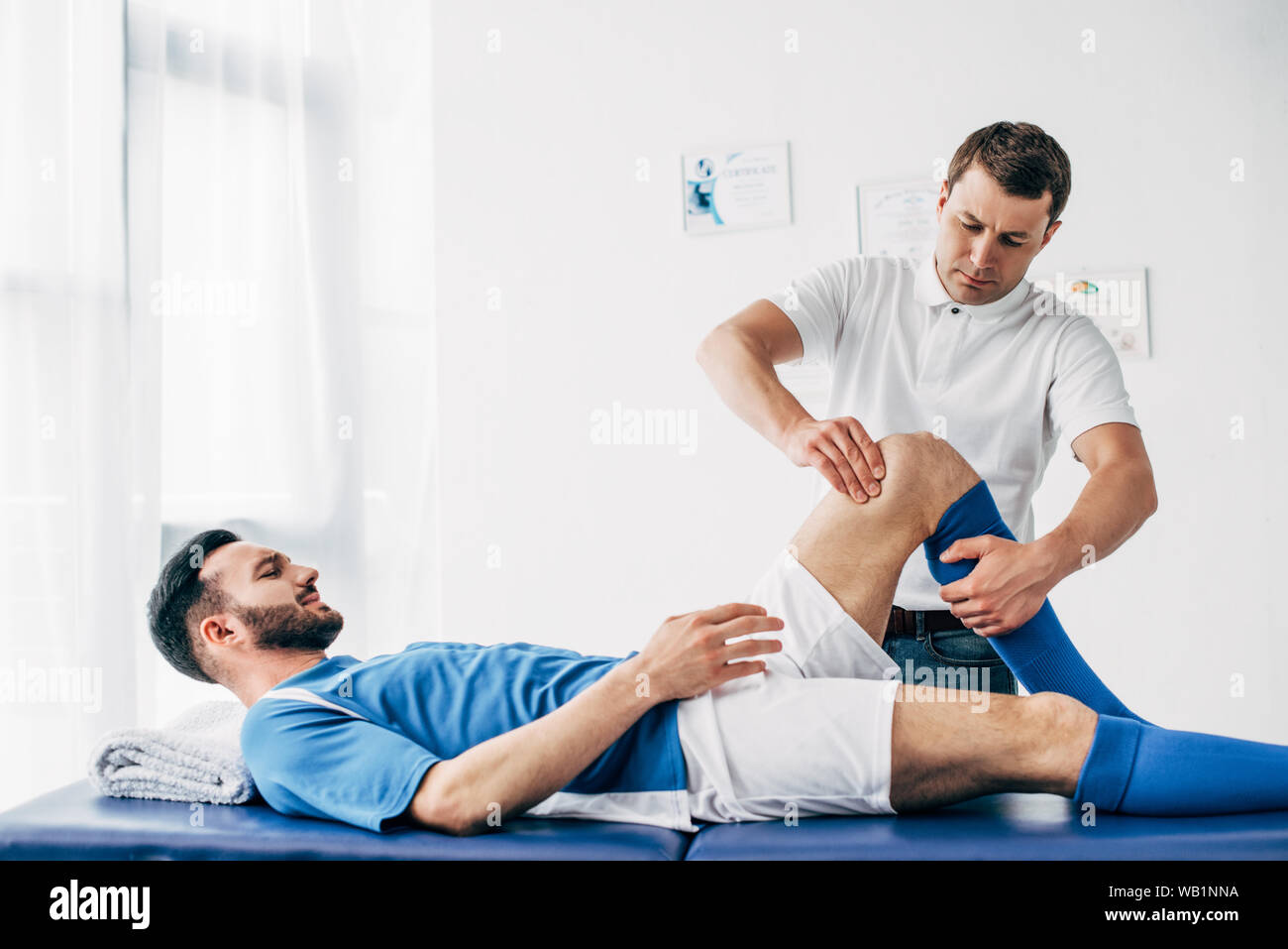 Physiotherapist massaging leg of football player lying on massage table in hospital Stock Photo