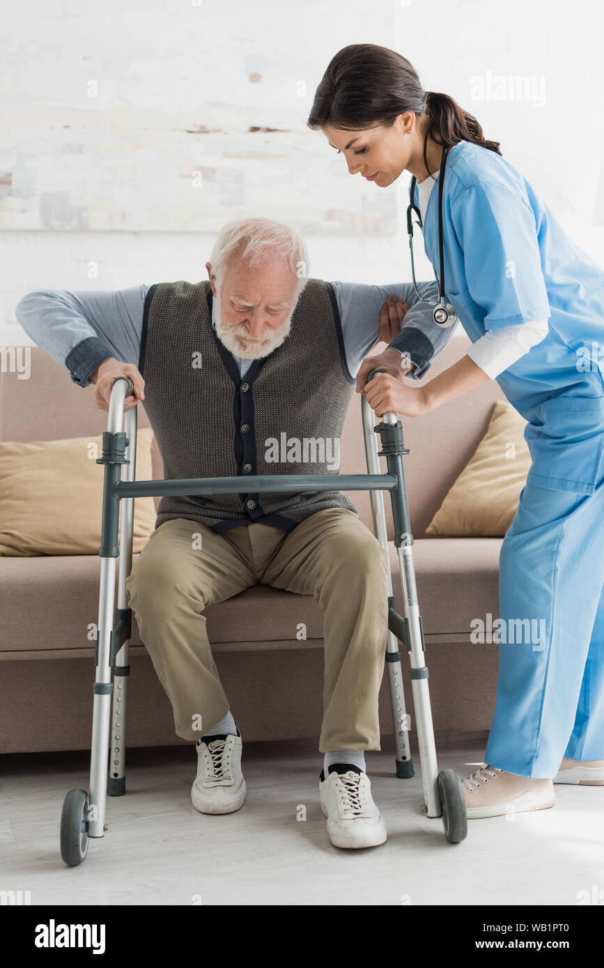 Nurse helping to retired man, getting up from sofa Stock Photo