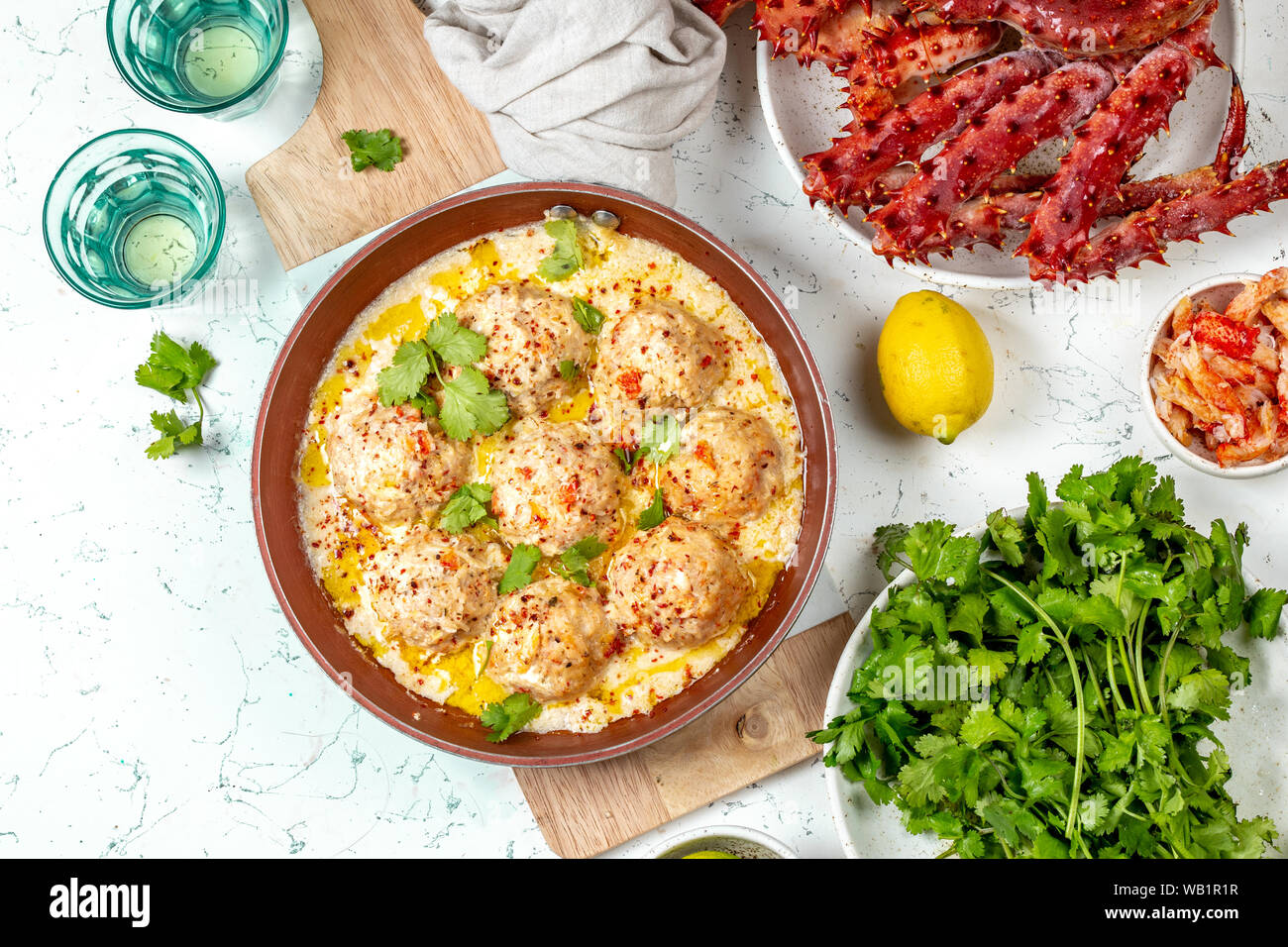 CRABMEATBALLS crab meatballs in white creamy sauce in red pan, whole king crab, cilantro, lemon and white wine on white background Stock Photo