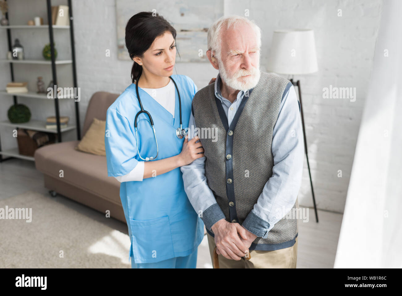 Doctor supporting of senior man, standing in bright light room, looking aside Stock Photo