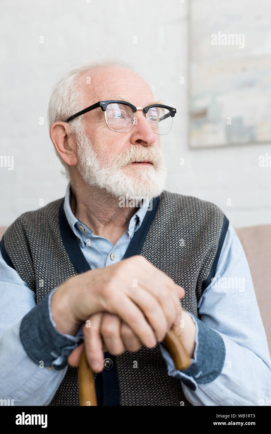 Calm and sad senior man with walking stick looking away, sitting in bright room Stock Photo