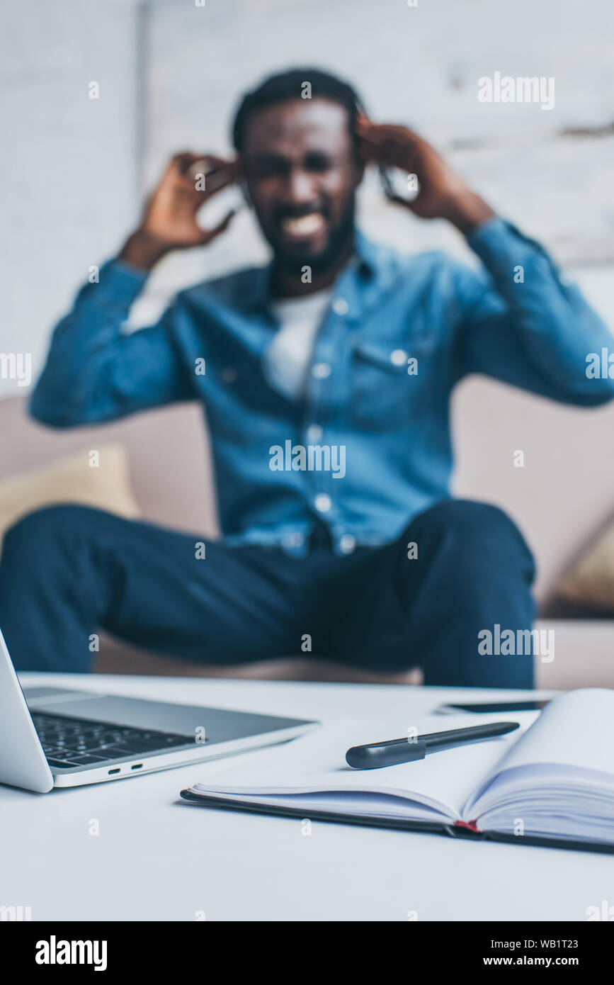 selective focus of african american man suffering from headache while sitting near table with laptop and notebook Stock Photo