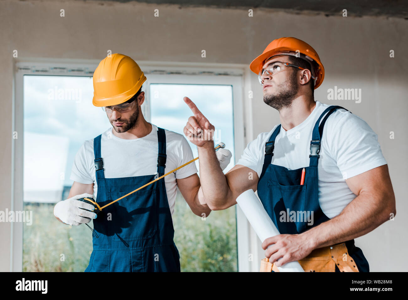 Pointing Near handsome handyman looking at measuring tape near coworker pointing with ...