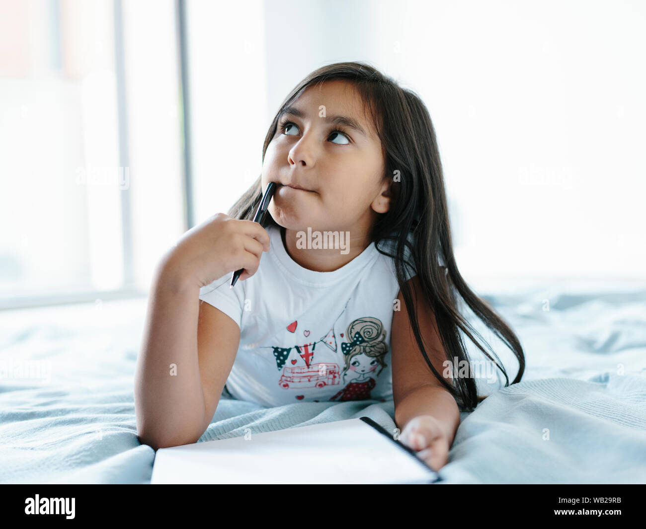 A 9year old girl is lying on a blanket on bed. She's writing on a notebook. She has long dark