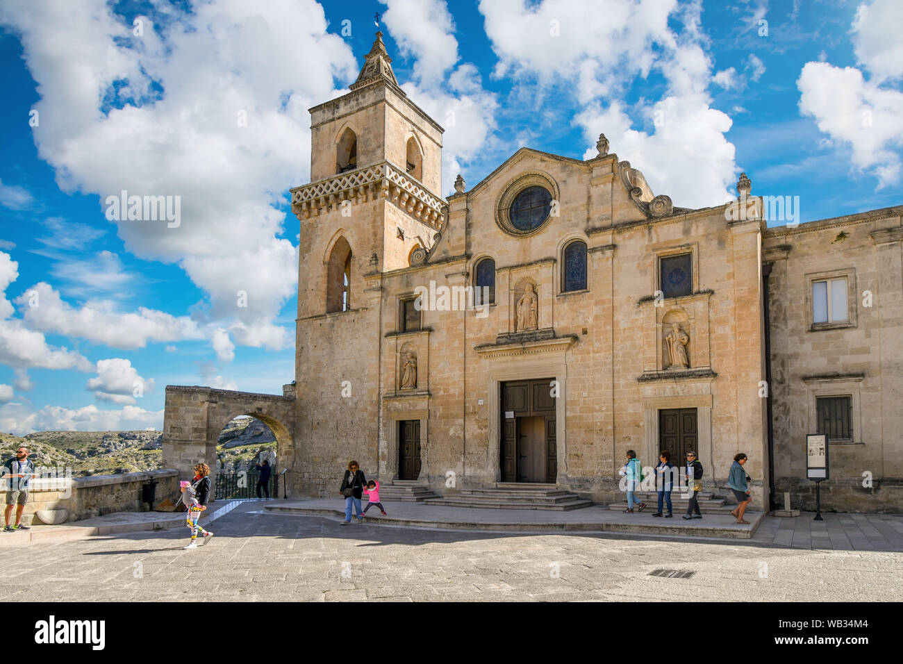 Tourists pass by the facade of the Church of San Pietro Caveoso with the ancient sassi caves in the distance on a summer day in Matera Italy Stock Photo