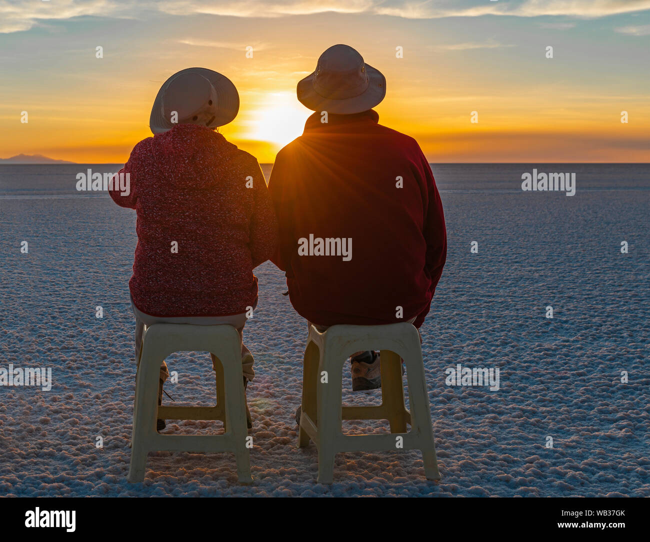 A couple of tourists sitting on benches and enjoying a sunset in the Uyuni salt flat desert with lens flare, Andes mountain range altiplano, Bolivia. Stock Photo