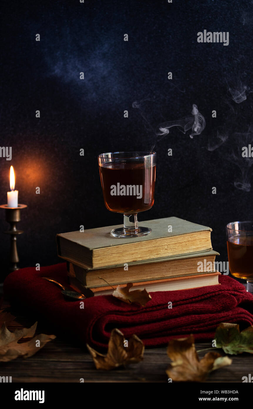 A cup of hot tea, books, a woolen knitted blanket, a candle, autumn leaves on a vintage wooden table. Autumn cozy mood. Low key. Copyspace. Stock Photo