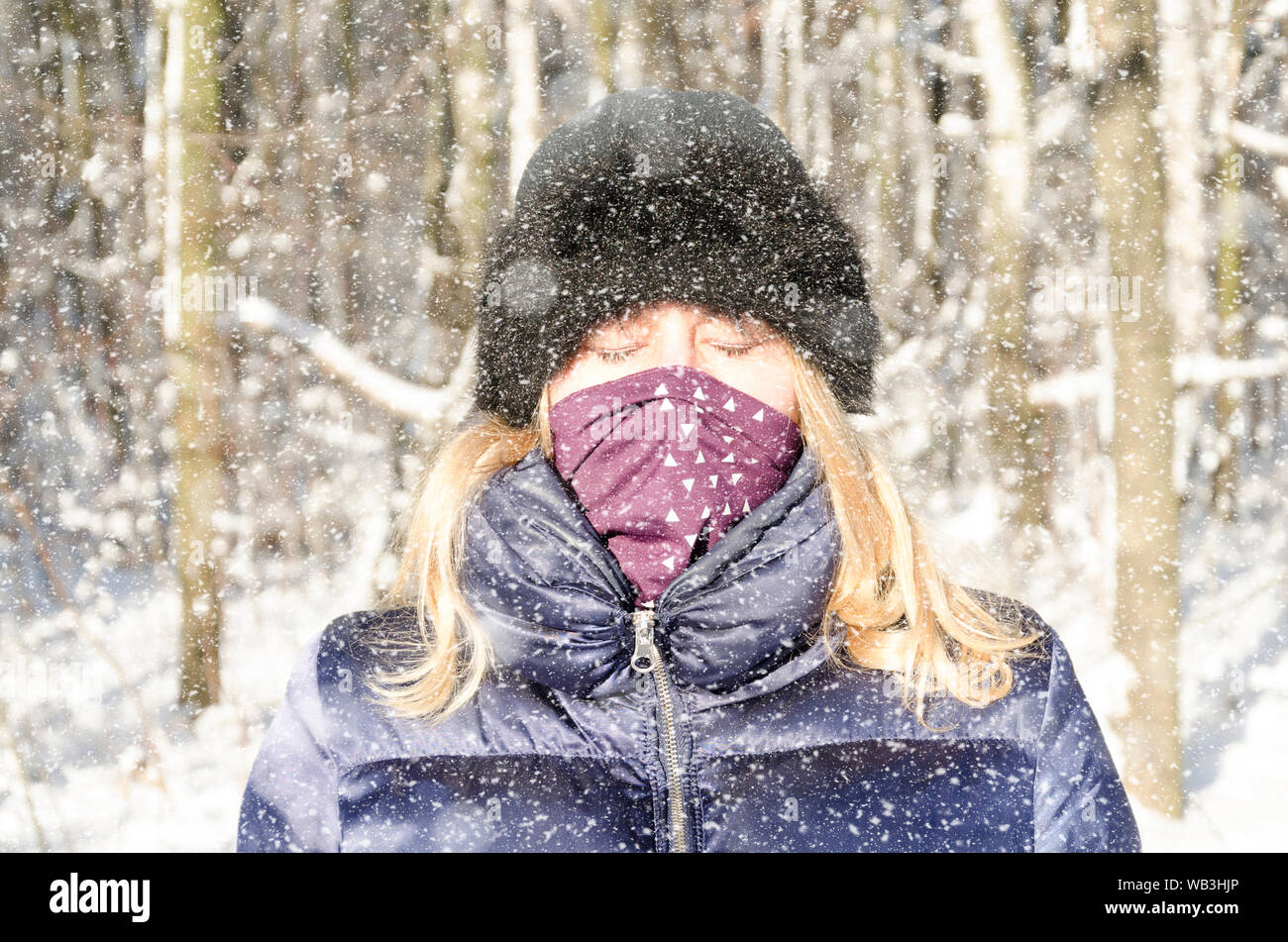 A blonde woman with her eyes closed and a scarf on her mouth enjoys the freezing cold of a snowy winter forest Stock Photo