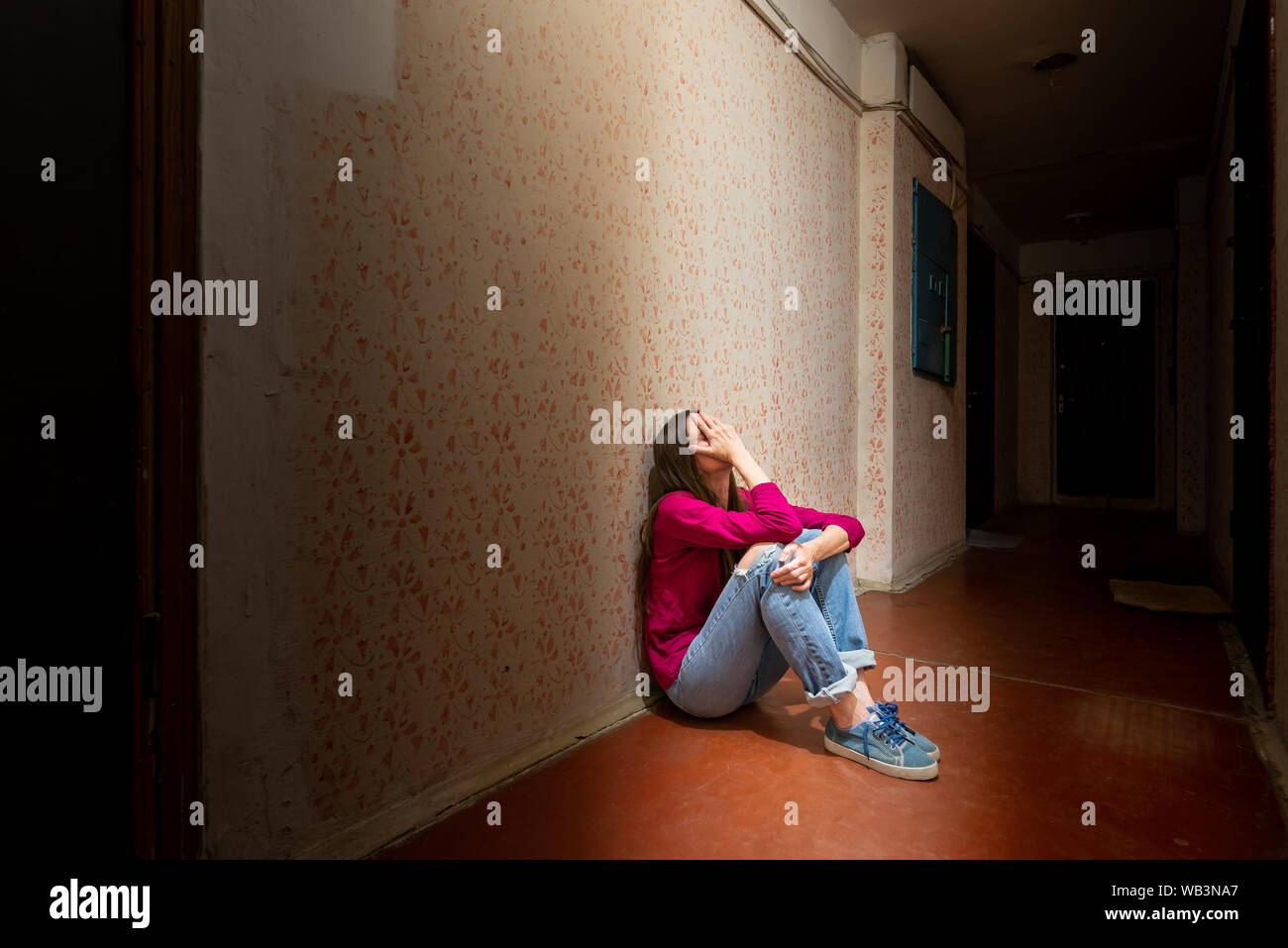 A sad and desperate woman sits in a dark corridor illuminated by a gloomy light. Her pain and her many problems pushed her into complete isolation. Hi Stock Photo