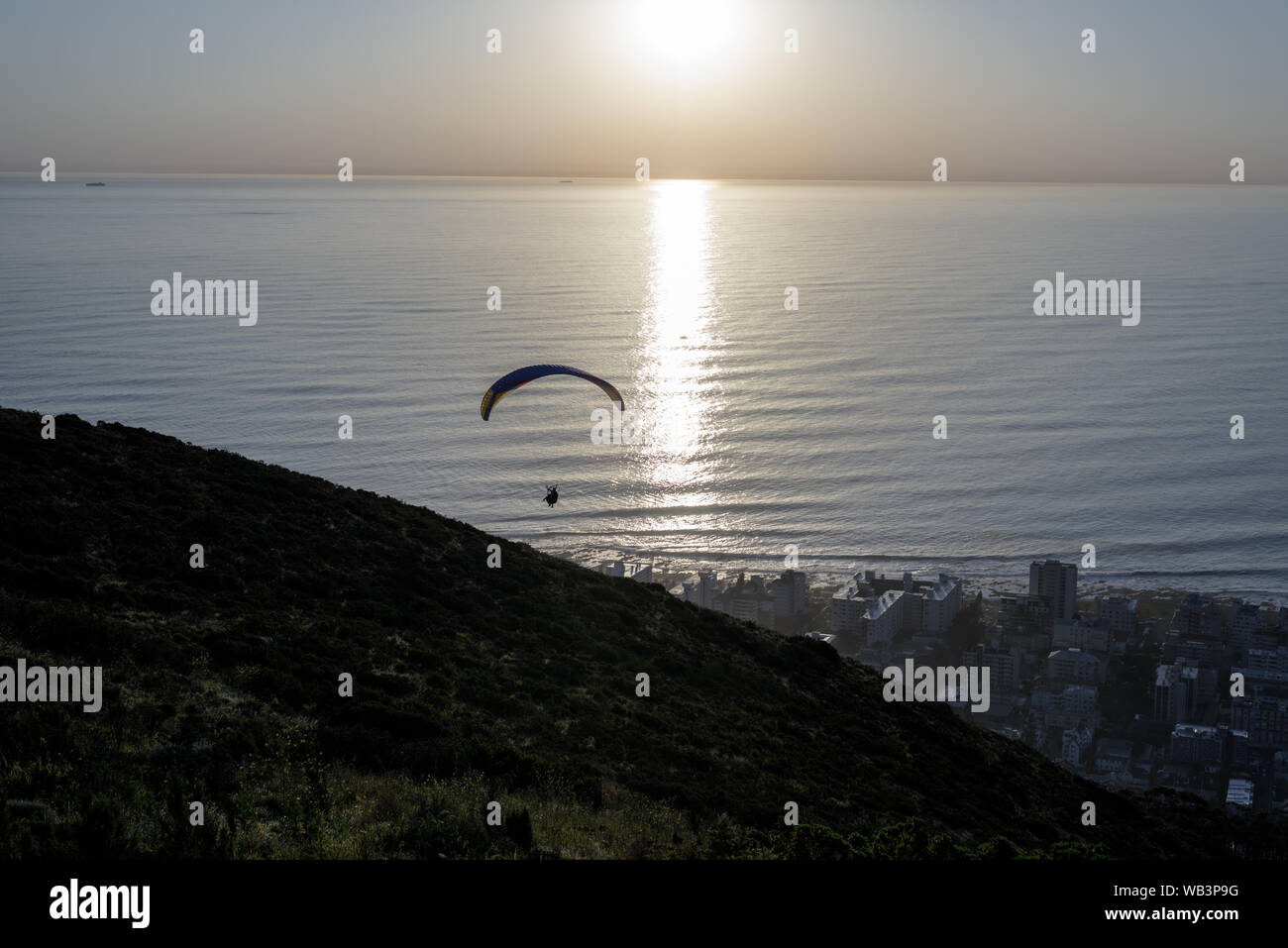 A paraglider after launching from Cape Town's Signal Hill at sunset above the Seapoint suburb on the South African Atlantic shoreline Stock Photo