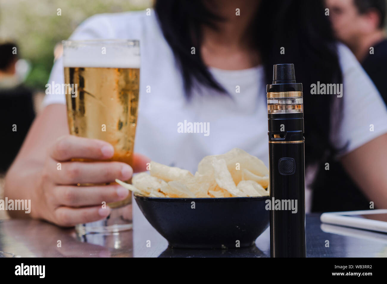 Woman drinking a beer on a terrace with electronic cigarette on the table and french fries Stock Photo
