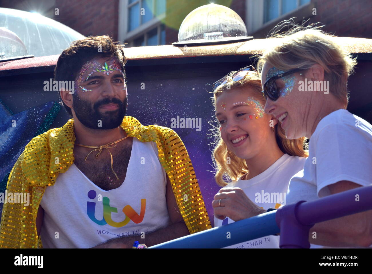 Coronation Street stars take part in the Manchester, uk, LGBT Pride Parade on 24th August, 2019. Charlie De Melo (left) who plays Imran Habeeb, wears a yellow cape as he talks to Sally Dynevor (right), who has played Sally Metcalfe in the soap since 1986, as her 24 year old daughter, Phoebe, stands between them. Stock Photo