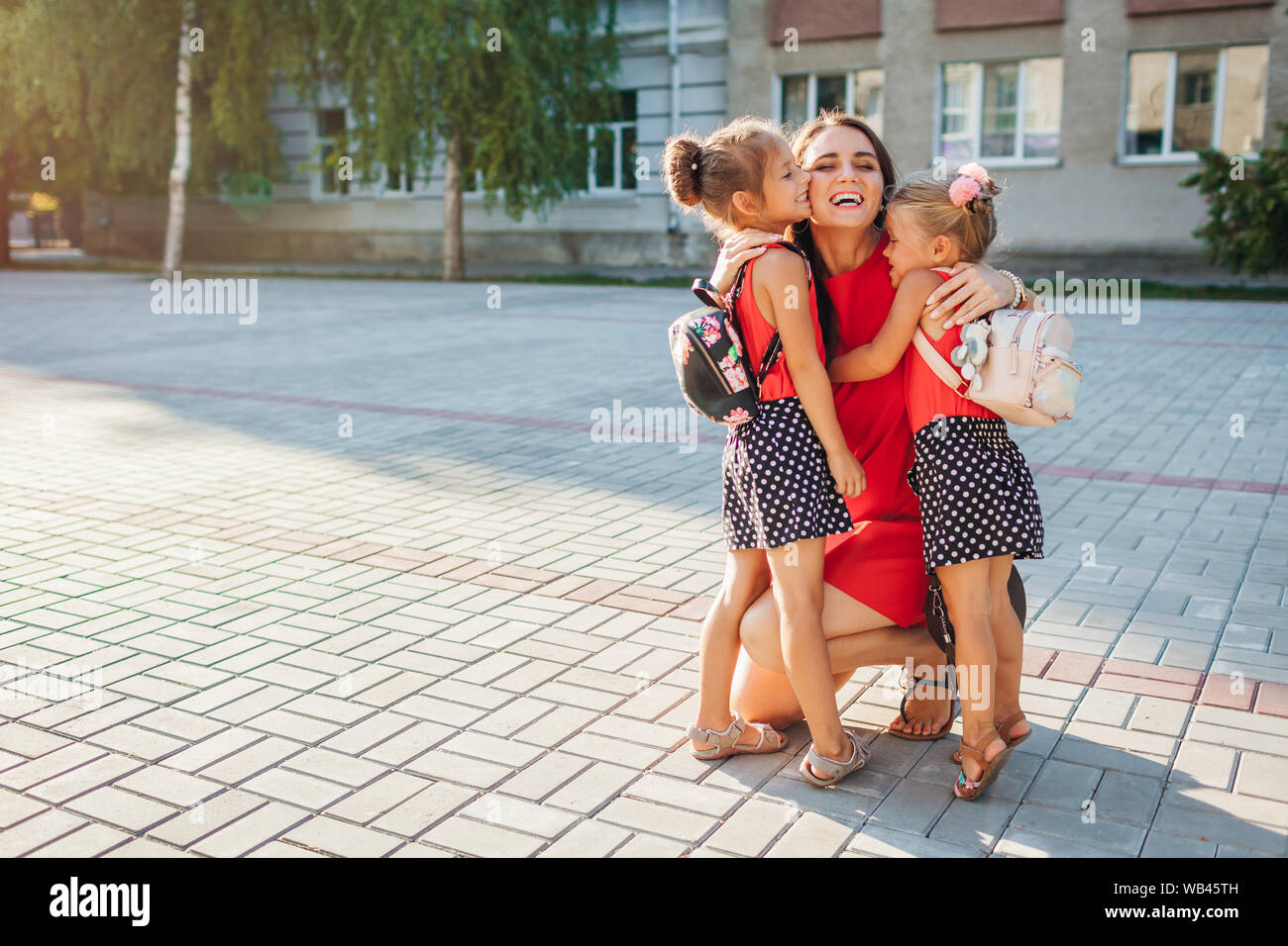 Happy mother meeting her kids daughters after classes outdoors primary school. Family hugging. Back to school concept Stock Photo