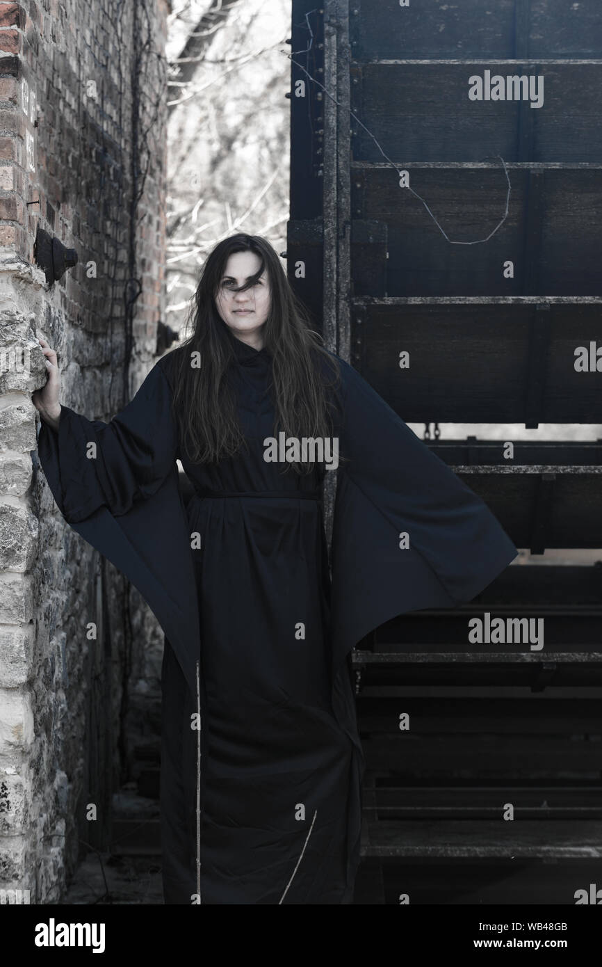 Young Woman with dark long hair in black robes in front of an Old Wooden Water Mill. Witches. Halloween concept. Witchcraft and Magic. Stock Photo