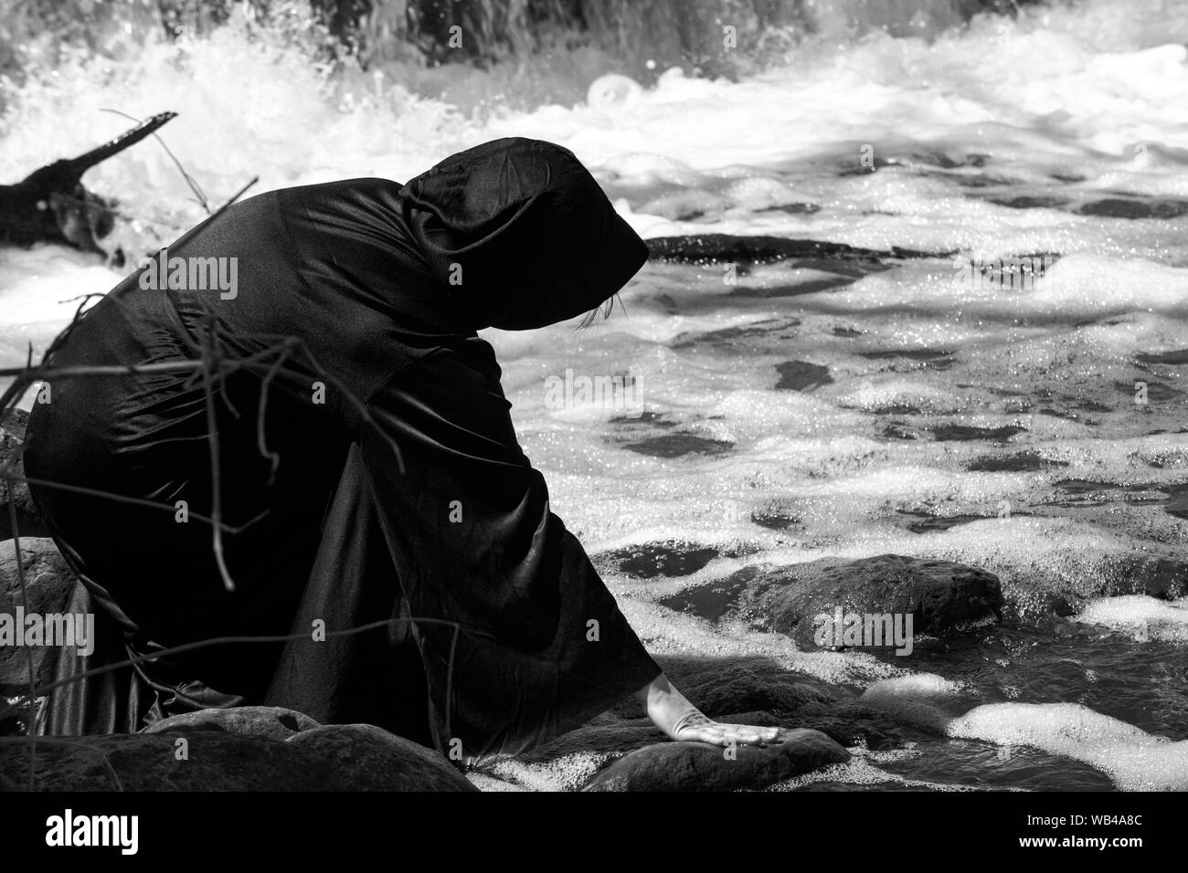 Hooded Woman in black robes in front of the Waterfall. Witches. Halloween and Gothic concept. Witchcraft and magic. Stock Photo