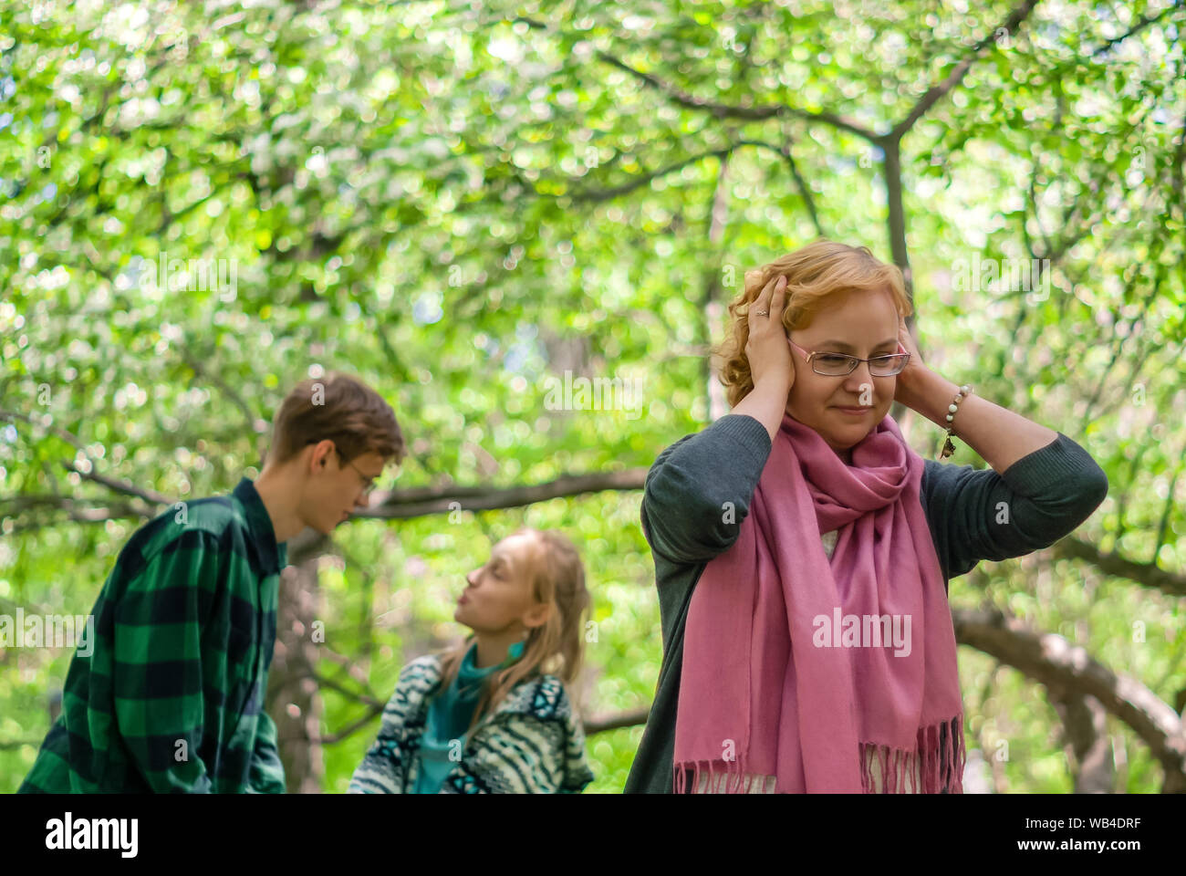 woman clamps her ears, seeking to enjoy the silence, while her two teenage children rustle in the background outdoor Stock Photo