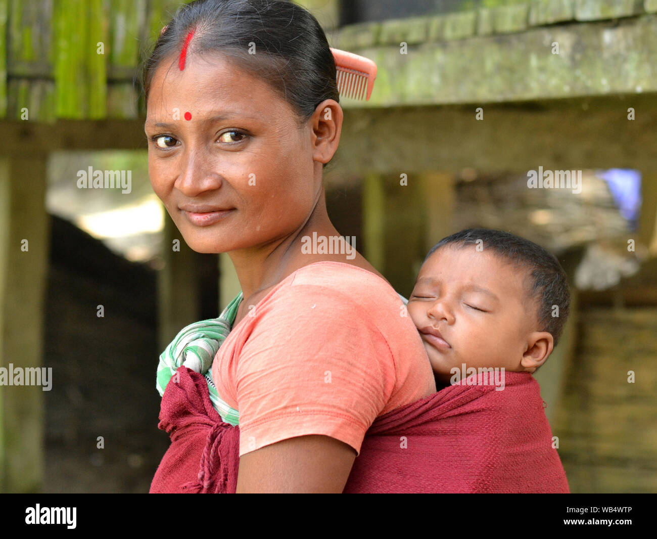 Married Indian Mishing tribal woman (red sindoor and bindi on forehead) from Majuli Island carries her sleeping baby boy in a traditional baby sling. Stock Photo