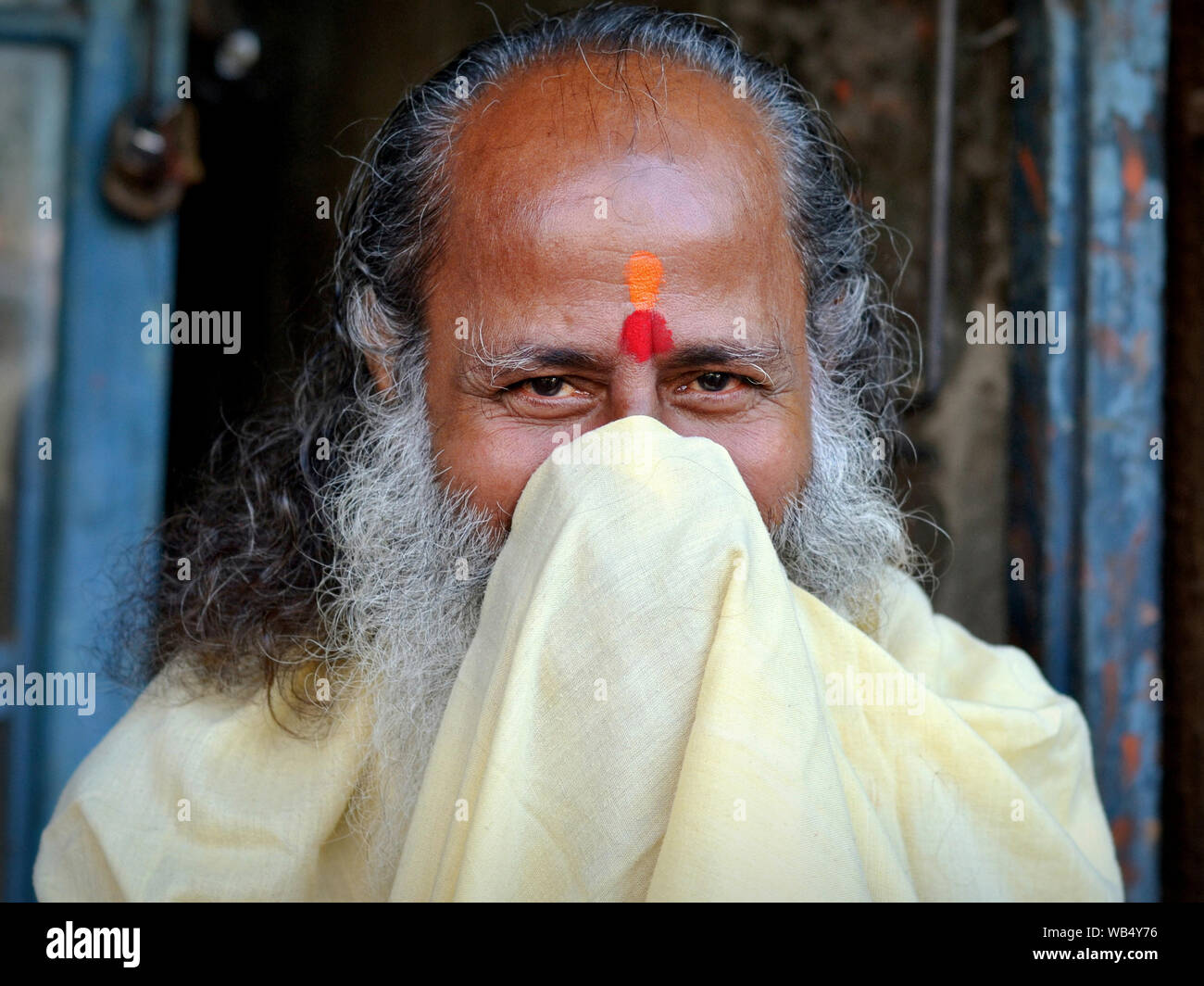 Smiling elderly Hindu priest (pujari) covers the lower part of his face with his robe and blow his nose with the priest's robe. Stock Photo
