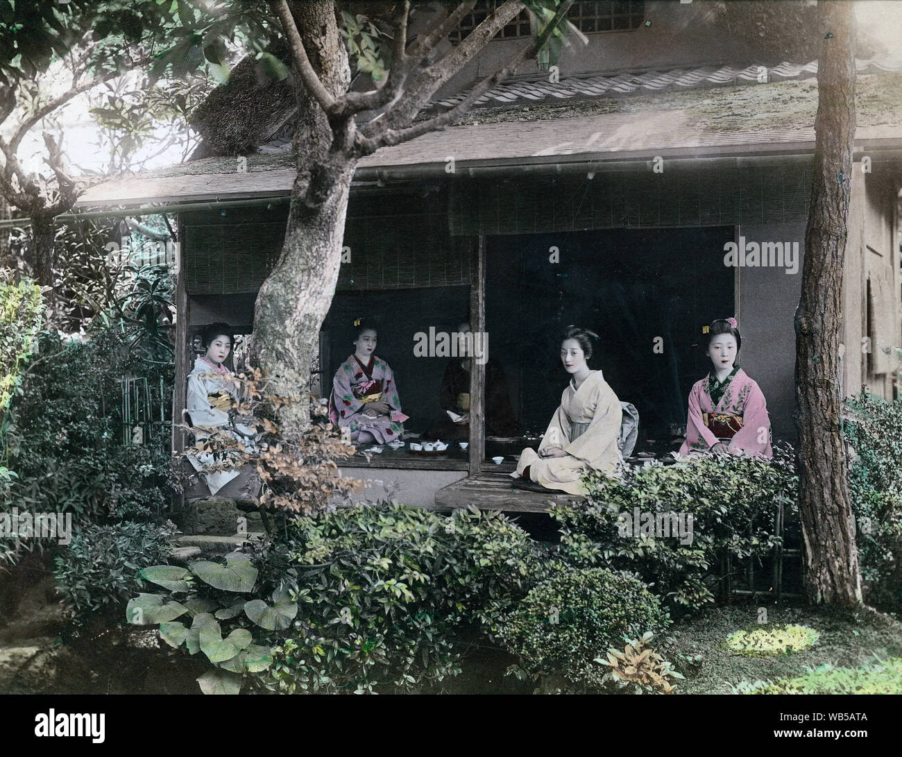 [ 1890s Japan - Japanese Women in Kimono ] —   Five women in kimono and traditional hairstyles are having tea in a cottage with thatched roof and Japanese garden. Two are sitting on the engawa (porch).  19th century vintage albumen photograph. Stock Photo