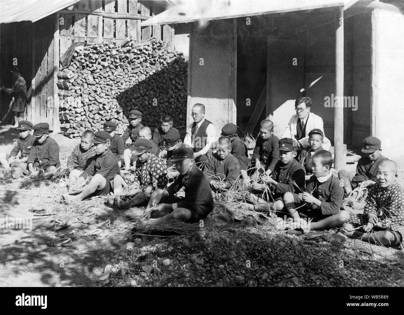 [ 1930s Japan - Japanese Elementary School Students ] —   Japanese male elementary school children in uniform sitting on the ground next to a farm building are making waraji (straw sandals). c1920s or 1930s.  20th century vintage gelatin silver print. Stock Photo