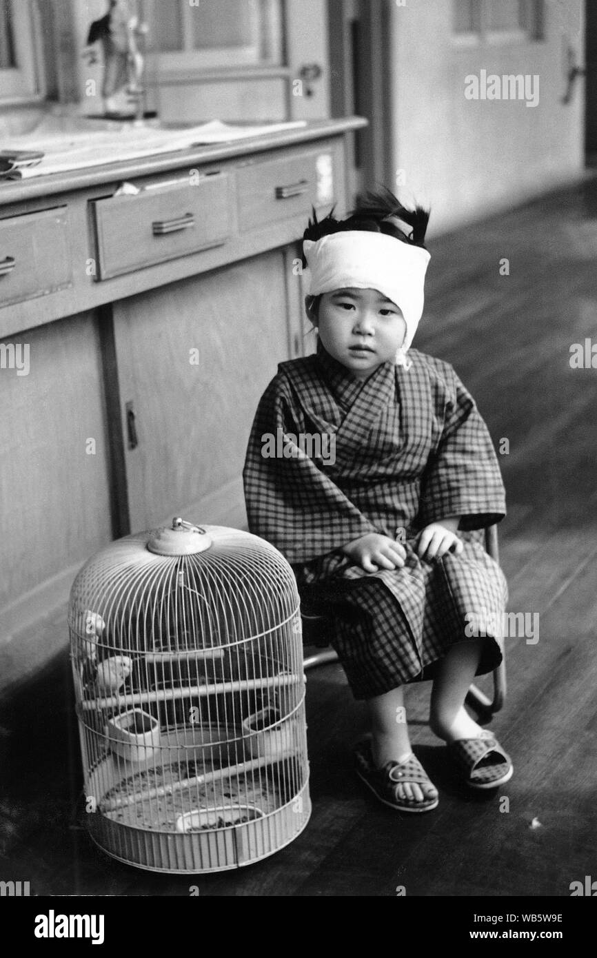 [ 1950s Japan - Japanese Girl with Bandage around Head ] —   A young girl with a bandage around her head waits at a hospital. Next to her stands a small cage with a bird. Late 1950s.  20th century vintage gelatin silver print. Stock Photo