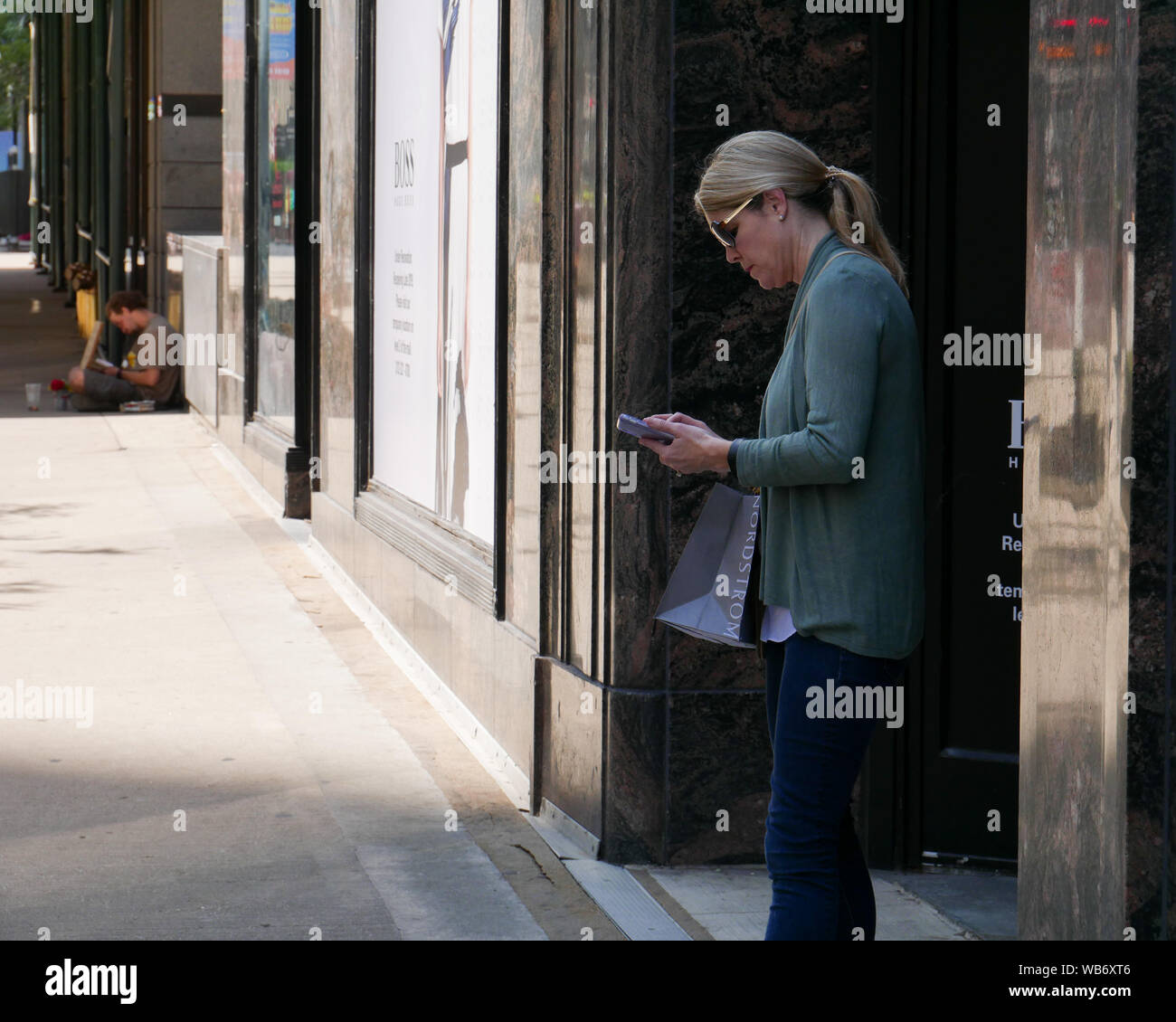 Woman with Nordstrom's bag checking phone. Homeless man in background. Chicago, Illinois. Stock Photo