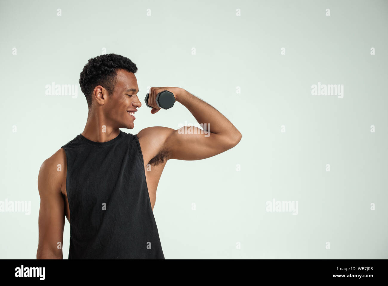 Making perfect body. Muscular young african man exercising with dumbbells and smiling while standing against grey background. Studio shot. Sport concept Stock Photo