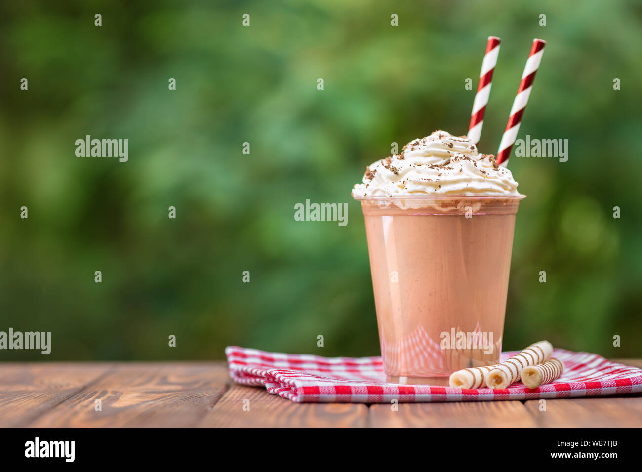 chocolate milkshake in disposable plastic cup with whipped cream and wafer rolls on wooden table outdoors Stock Photo