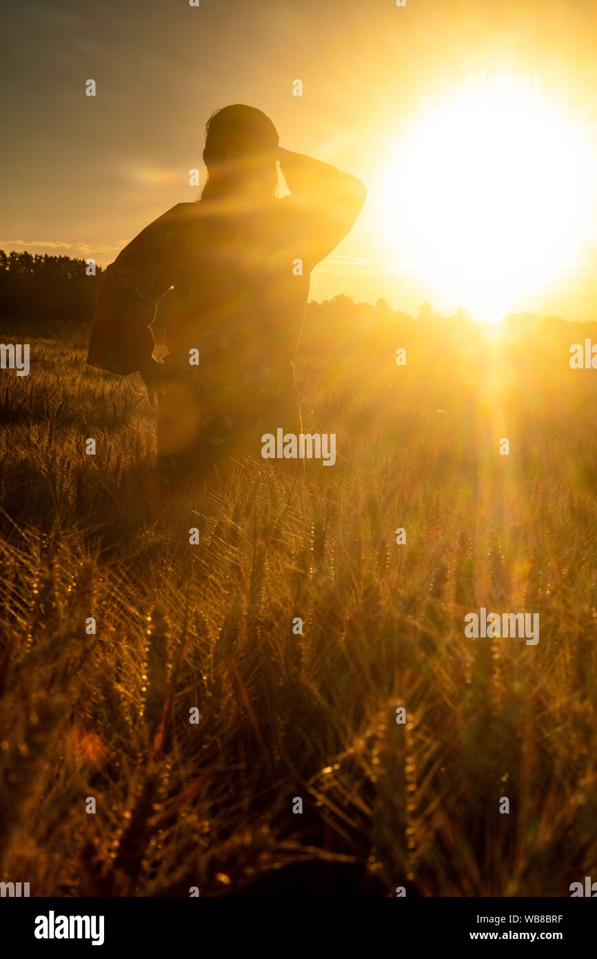 African woman in traditional clothes standing, looking, hand to eyes, in field of barley or wheat crops at sunset or sunrise Stock Photo