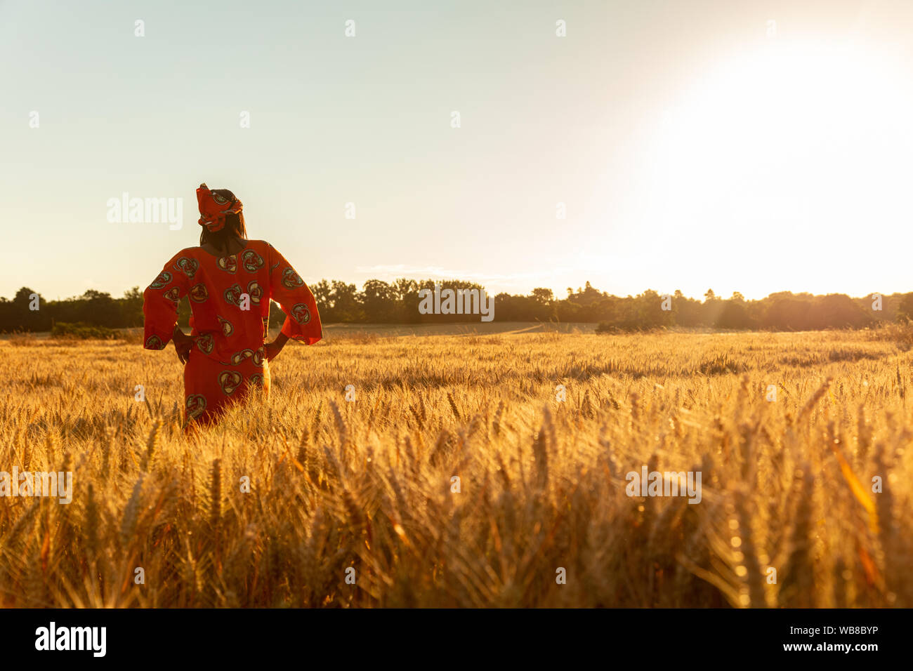 African woman in traditional clothes standing, looking, hand to eyes, in field of barley or wheat crops at sunset or sunrise Stock Photo