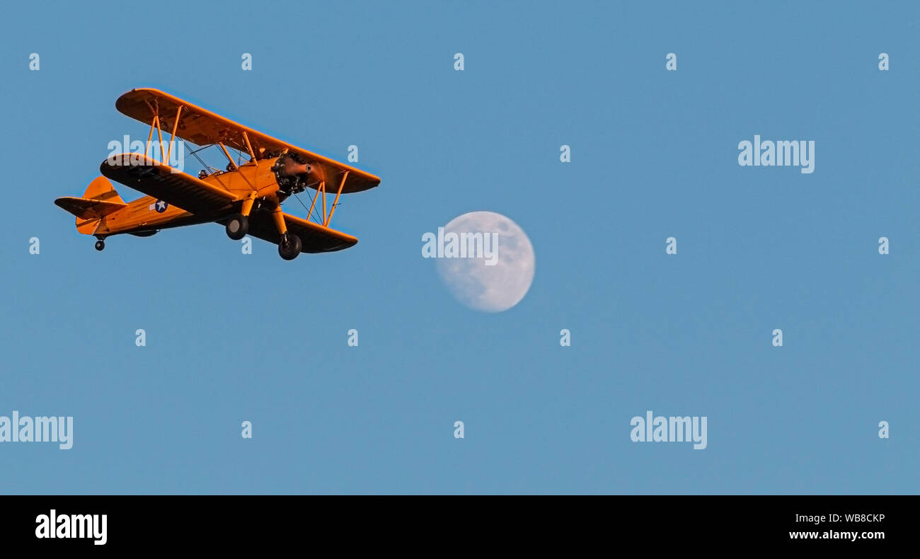 An antique american bi plane flying overhead at dusk with full moon and a blue sky. Stock Photo
