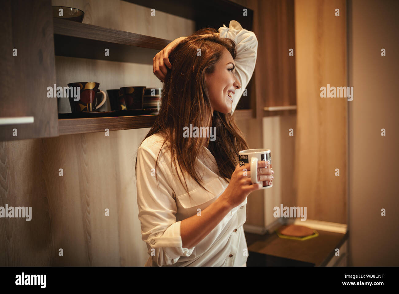 A young happy smiling woman is drinking morning coffee sitting in the kitchen after awakening. Stock Photo