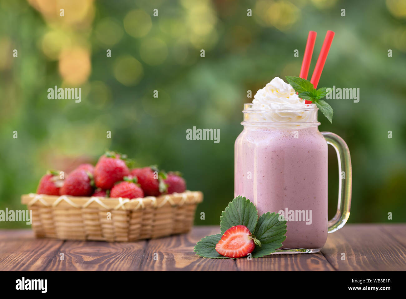 strawberry milkshake in mason jar with whipped cream and fresh ripe berries in basket on wooden table outdoors Stock Photo