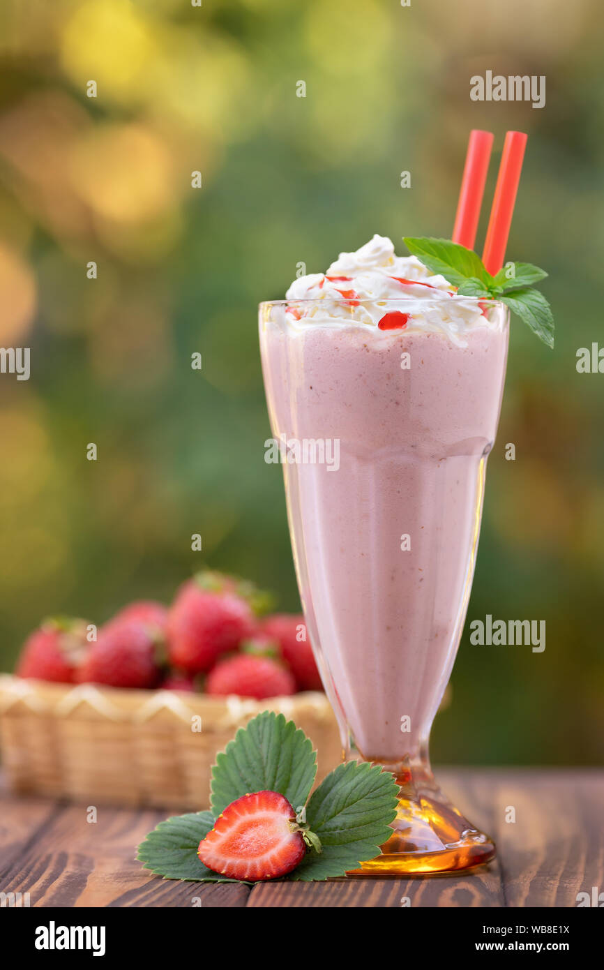 strawberry milkshake in glass with whipped cream and fresh ripe berries in basket on wooden table outdoors Stock Photo
