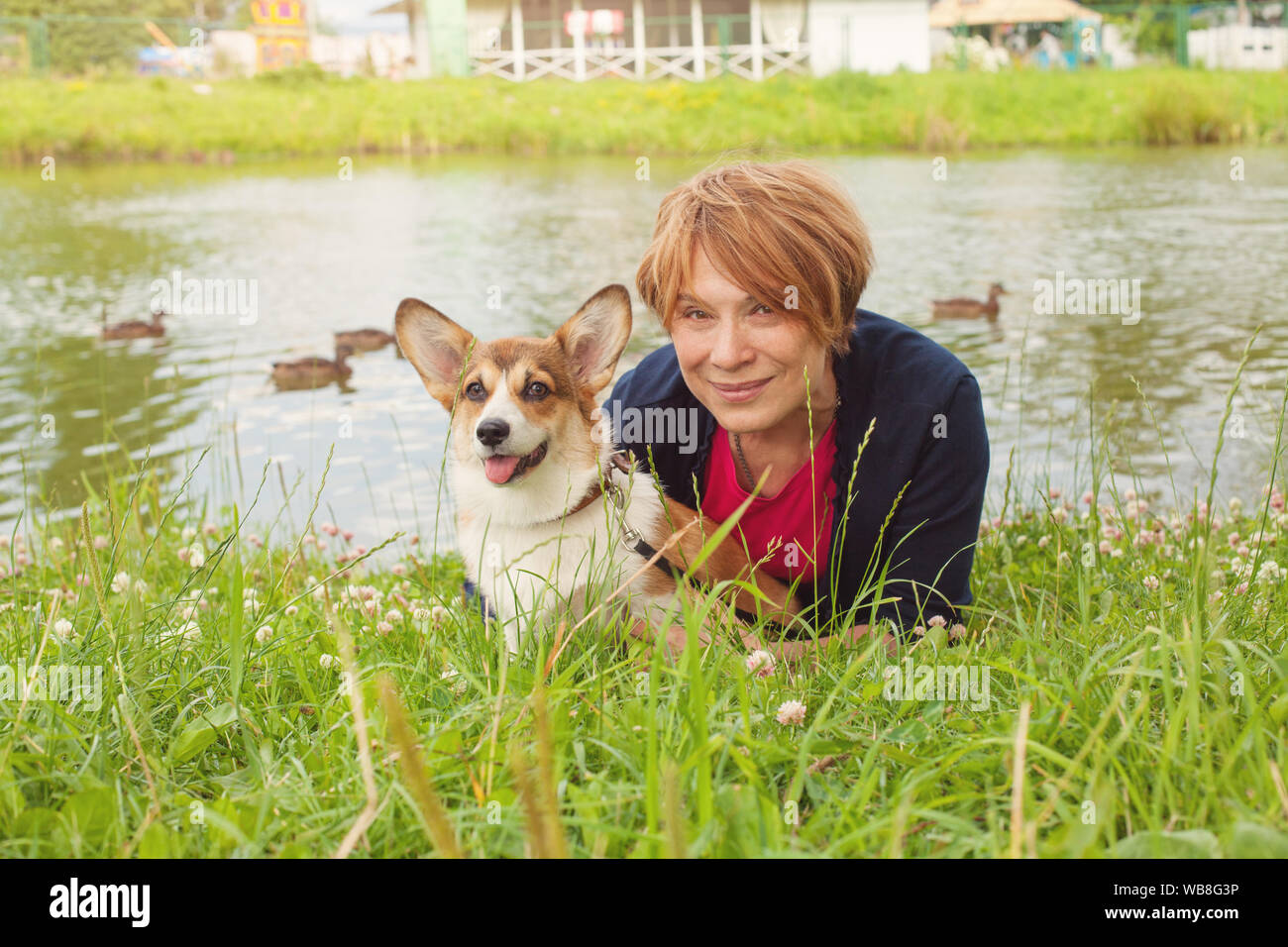 Pretty senior woman lying on green grass and hugging her corgi dog Stock Photo