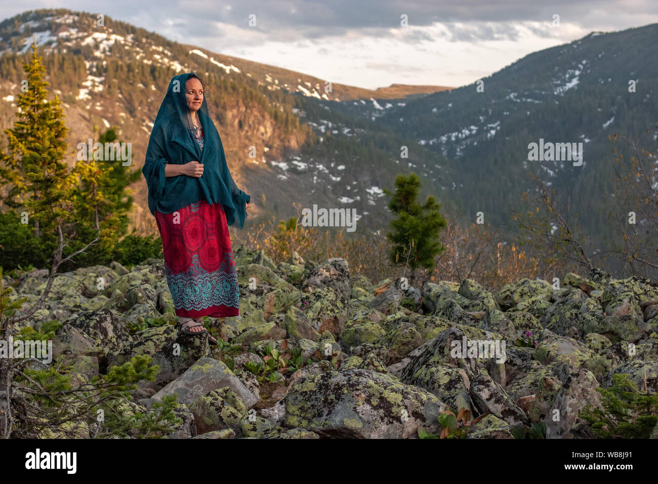 Beautiful cute girl in a dress and a scarf stands on large stones in the mountains. Against the background of mountains with snow overgrown with fores Stock Photo