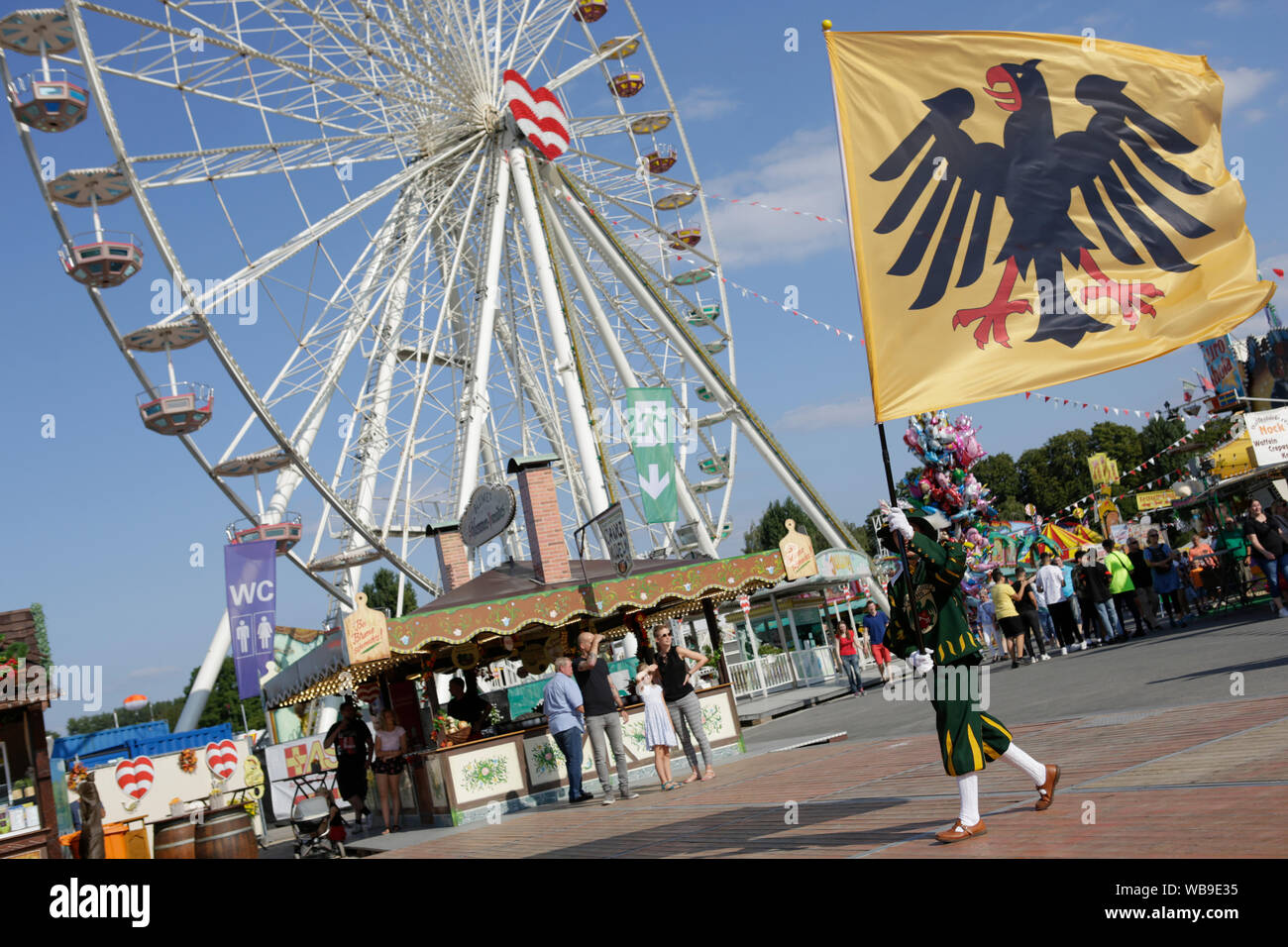 Worms, Germany. 24th August 2019. A colour guard from the Fanfare Corps of the city of Worms marches past a Ferris wheel. The largest wine and funfair along the river Rhine, the Backfischfest started in Worms with the traditional handing over of power from the Lord Mayor to the mayor of the fishermen’s lea. The ceremony was framed by dances and music. Stock Photo