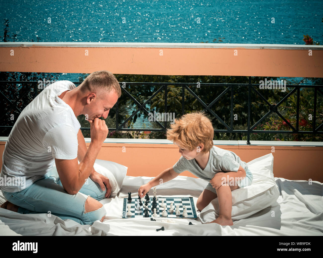 Father and little son play chess on balcony, nature and sea on background. Dad with child play intellectual game. Smart toddler concept. Parent play c Stock Photo