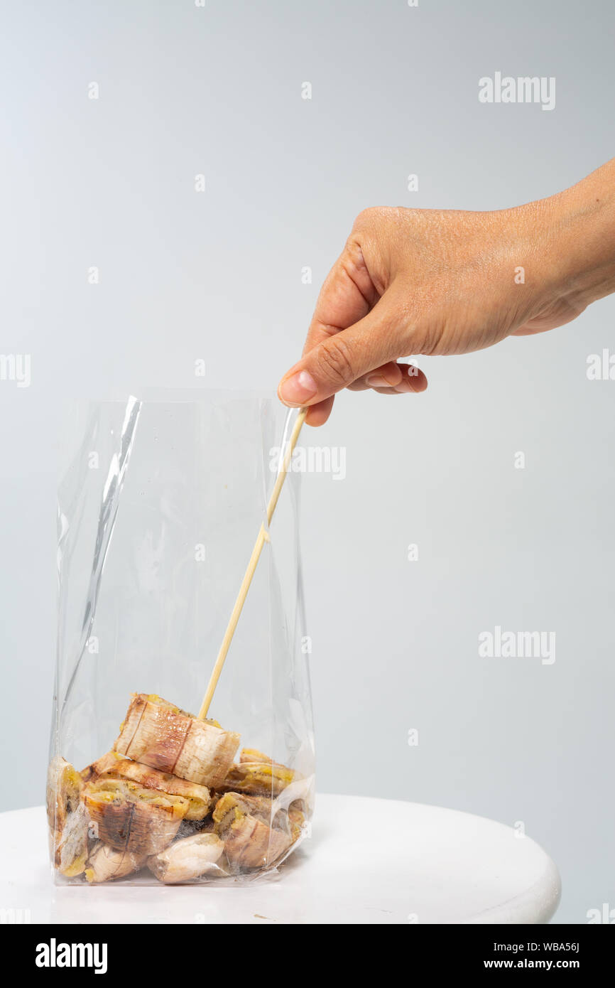 Woman's right hand using skewer pick up  grilled banana from plastic bag on grey background, Close up shot, Selective focus, Thai traditional street f Stock Photo