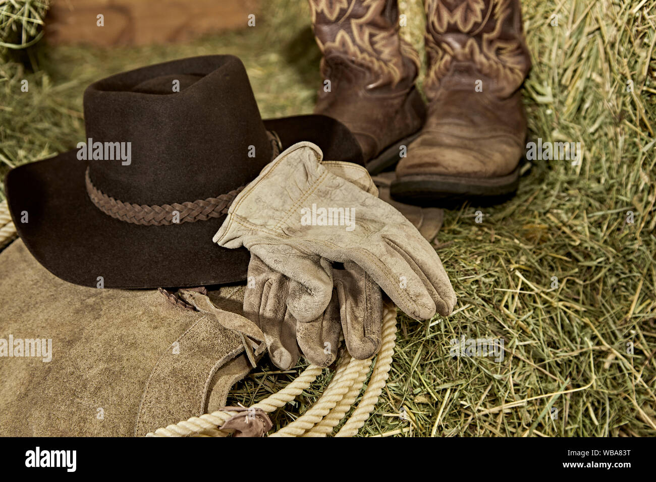 Western Cowboy hat with cowboy boots, leather gloves, leather chaps and a roper's rope on hay in a barn Stock Photo